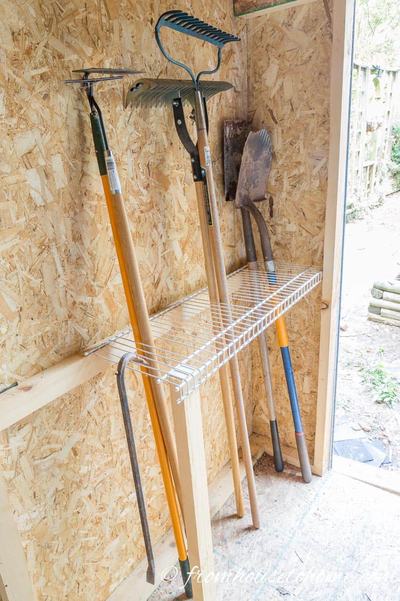 Hoes, rakes and shovels stored in a wire shelf attached to the shed wall