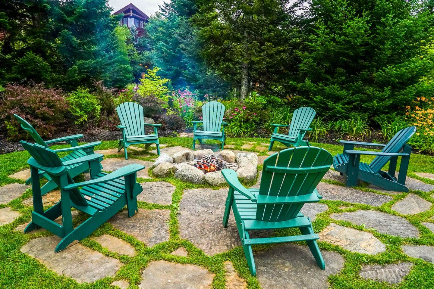 Firepit made with large rocks in the middle of a flagstone patio with 7 green adirondack chairs surrounding it