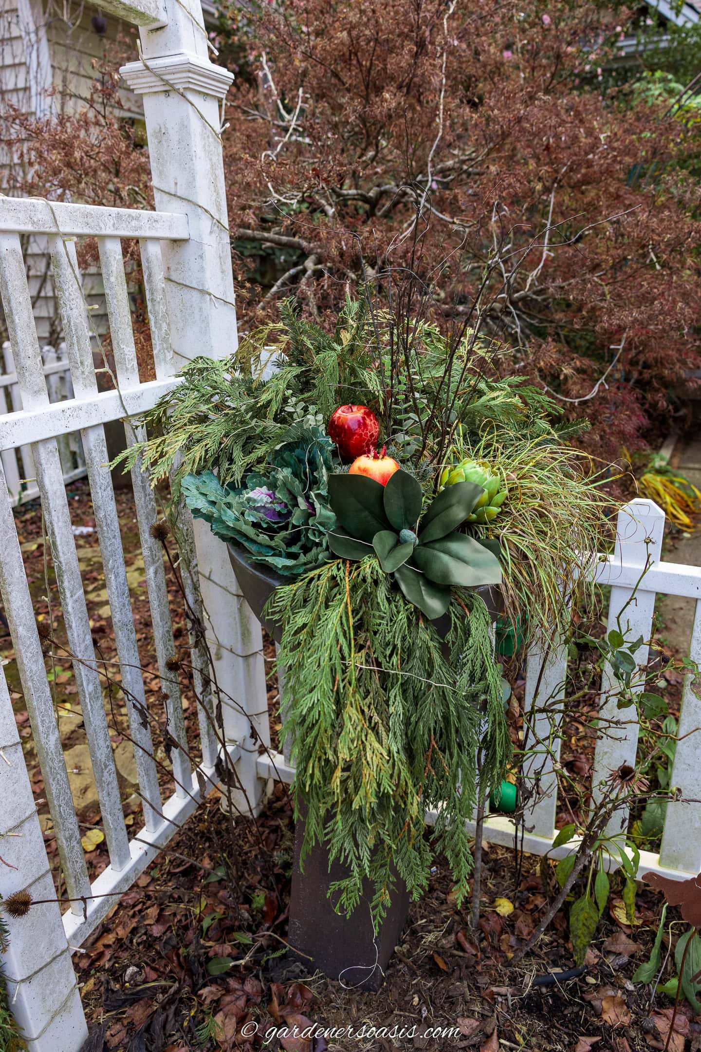 a modern tall winter planter made with cedar boughs, fir branches, ornamental cabbage, magnolia branch, curly branches and artificial fruit