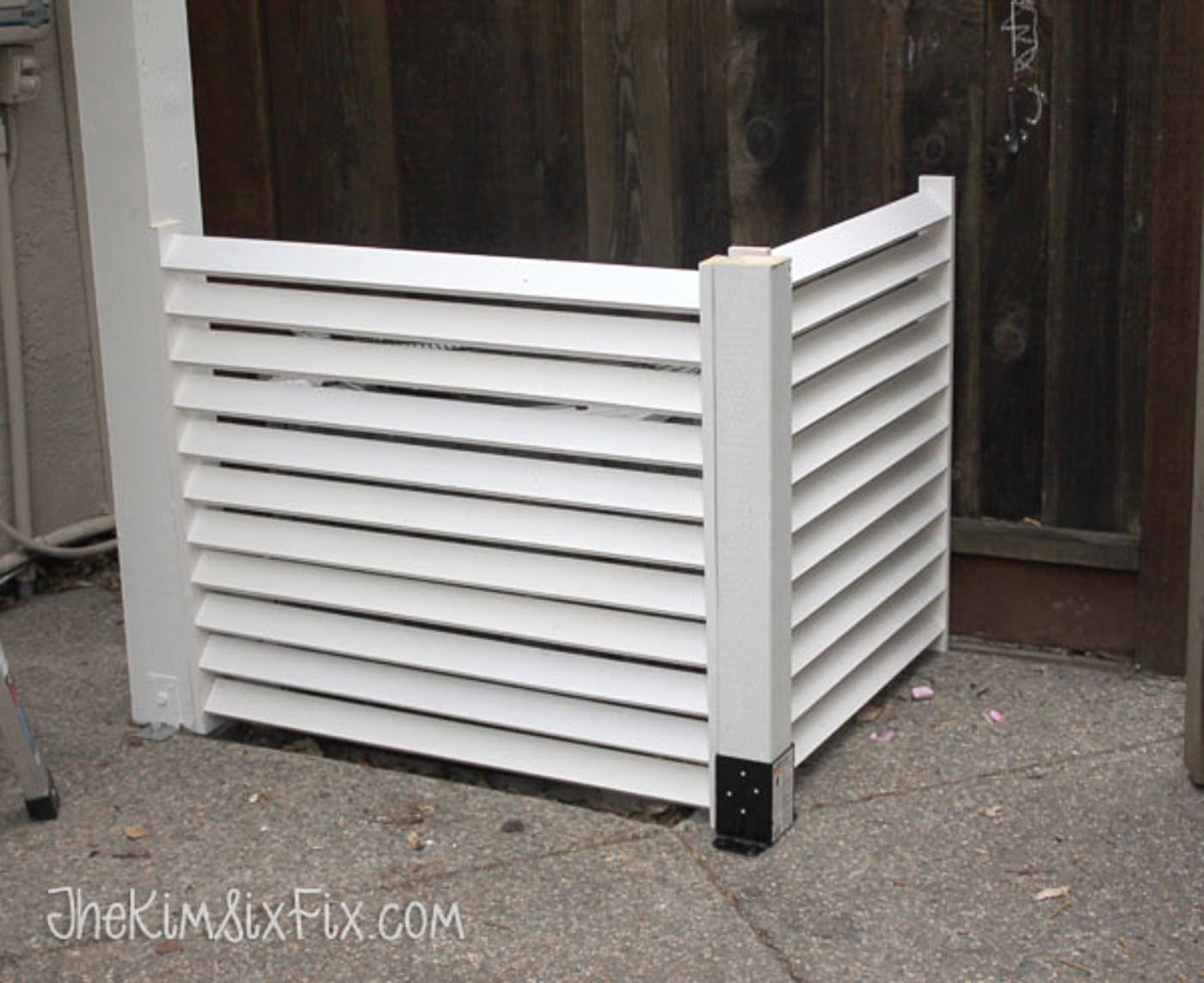 White wooden slatted enclosure covering an outdoor unit, placed on a concrete surface next to a wooden fence.