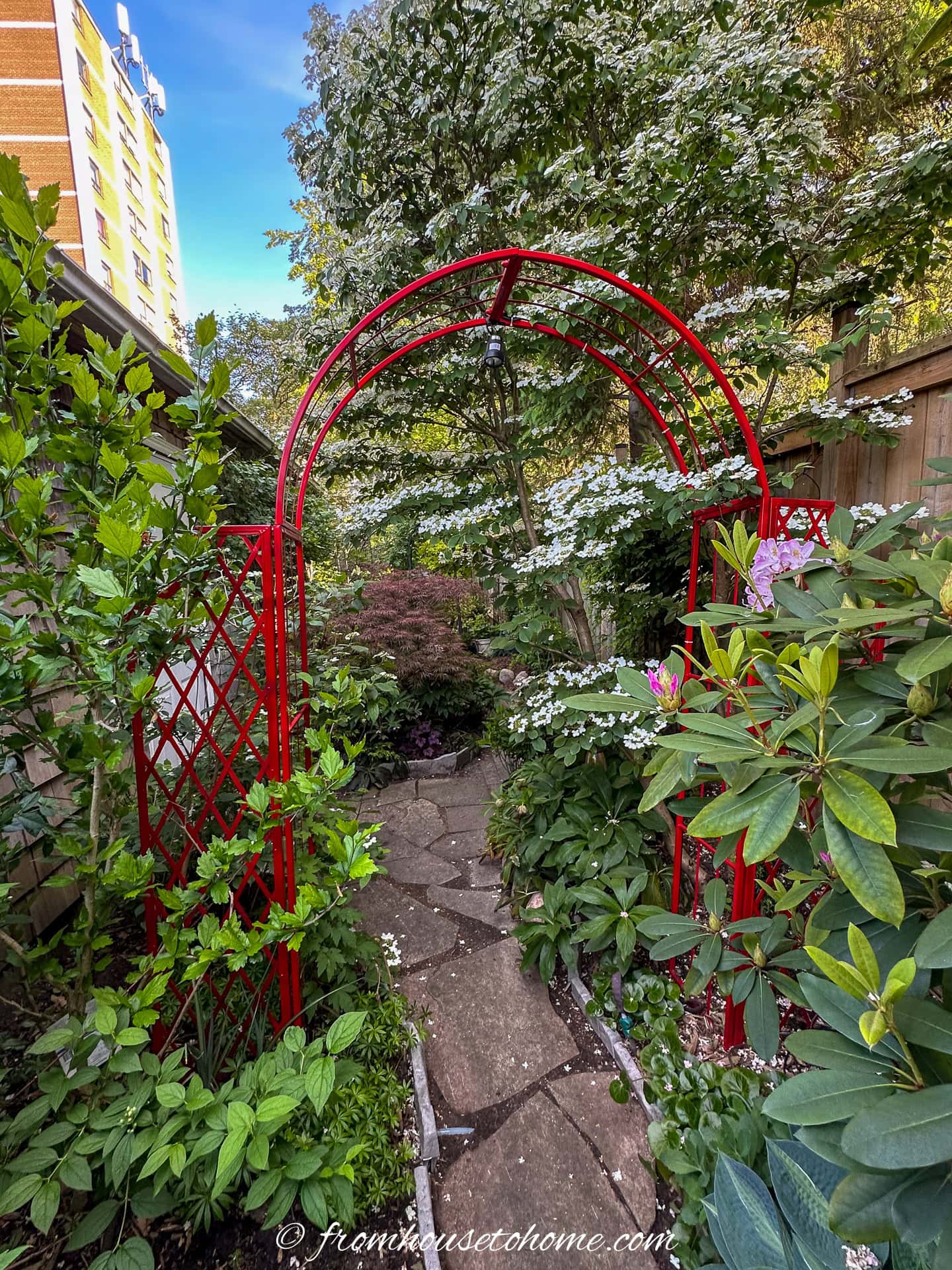 Garden path with stone steps leading through a red metal archway, surrounded by green plants and flowers, with a building visible in the background.