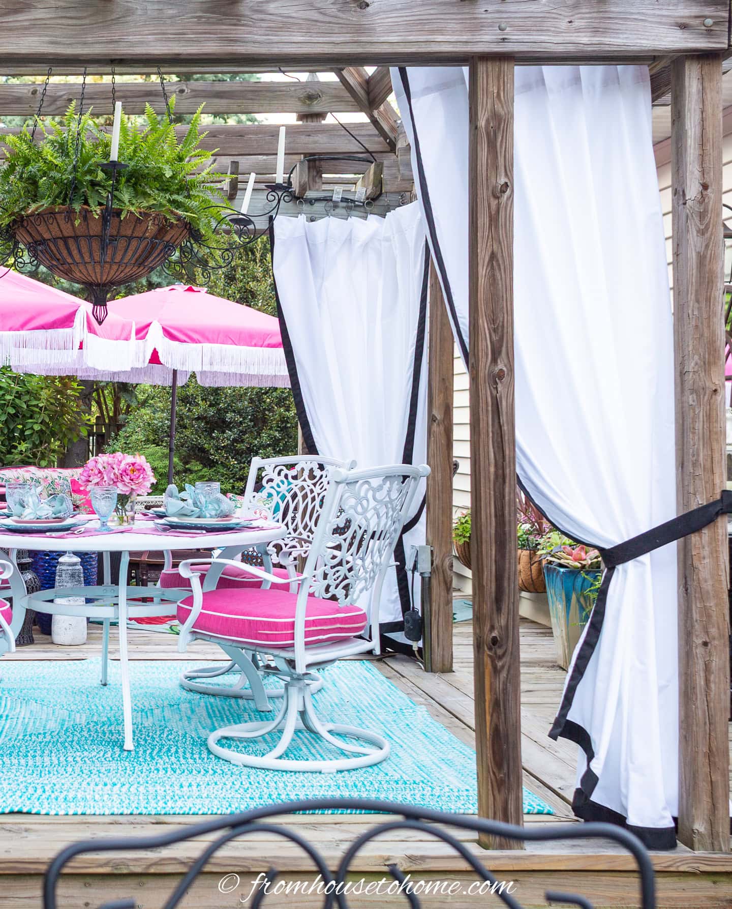 A patio with a white and pink umbrella, white table and chairs with pink cushions, a blue rug, potted plants, and white curtains tied to a wooden pergola.