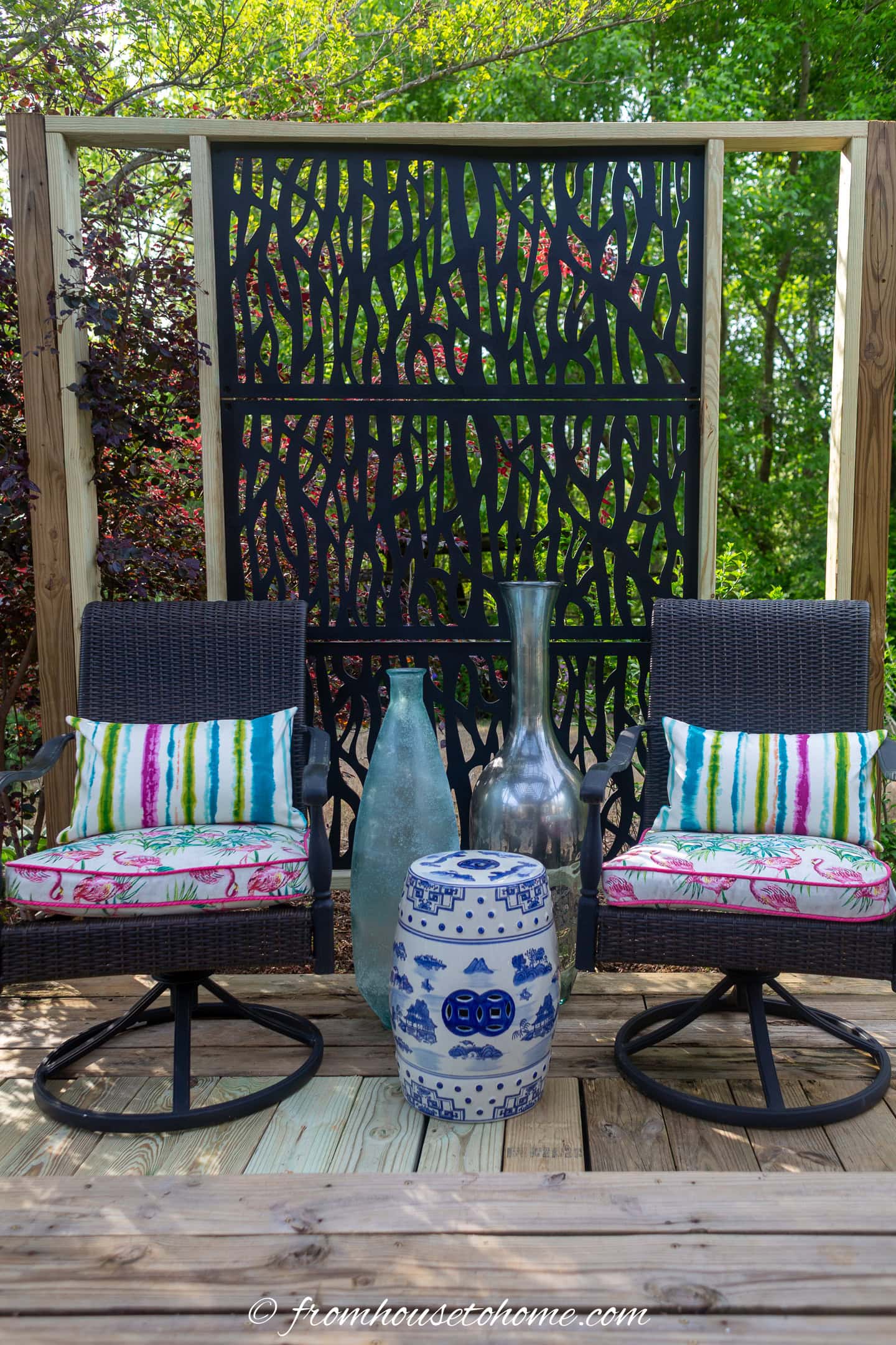 Two wicker chairs with colorful cushions sit on a wooden deck, separated by a decorative ceramic stool and tall vases, with a patterned privacy screen behind them.
