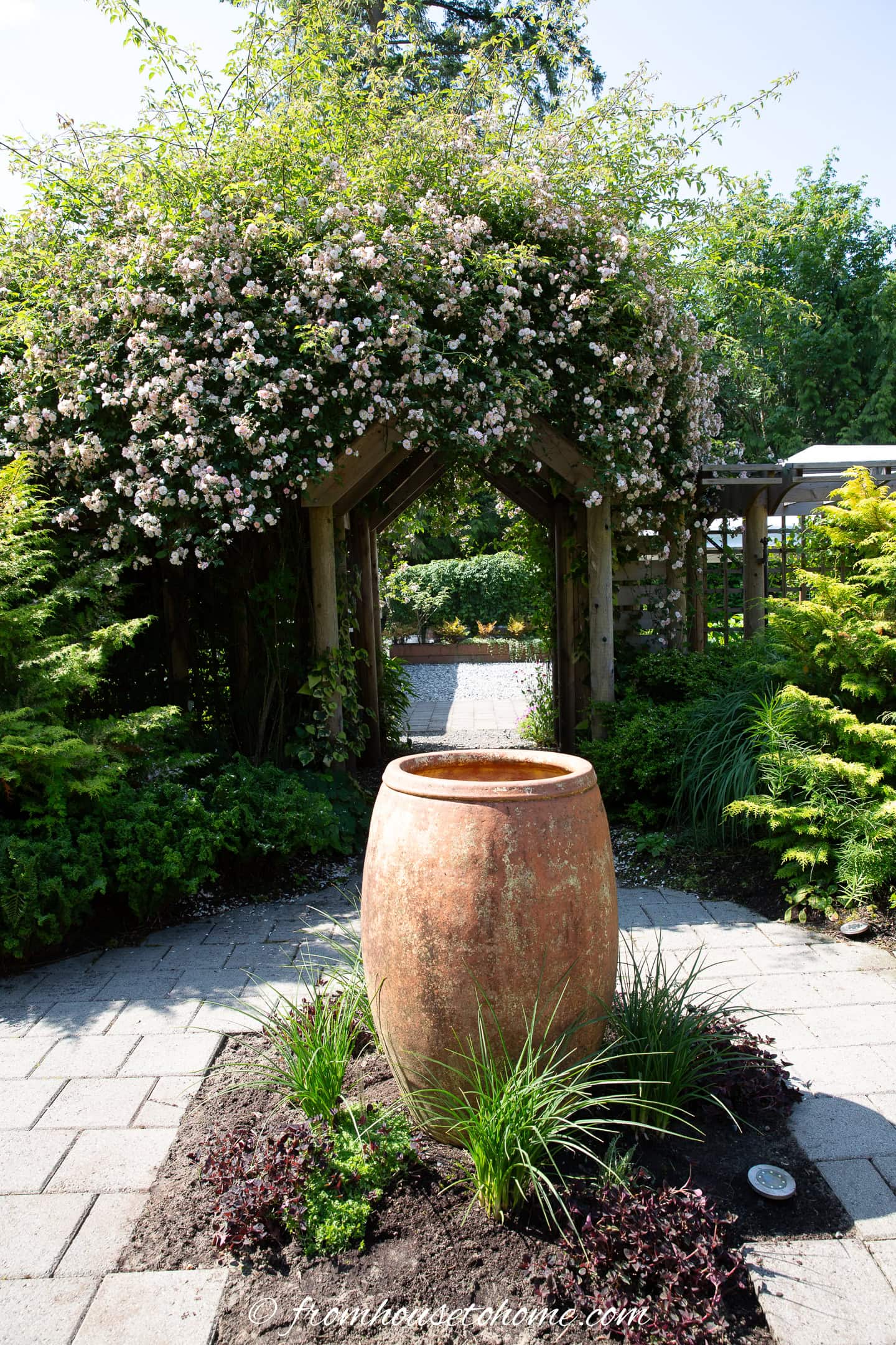 A large terracotta urn sits on a garden path, with a wooden archway covered in blooming flowers and greenery in the background.