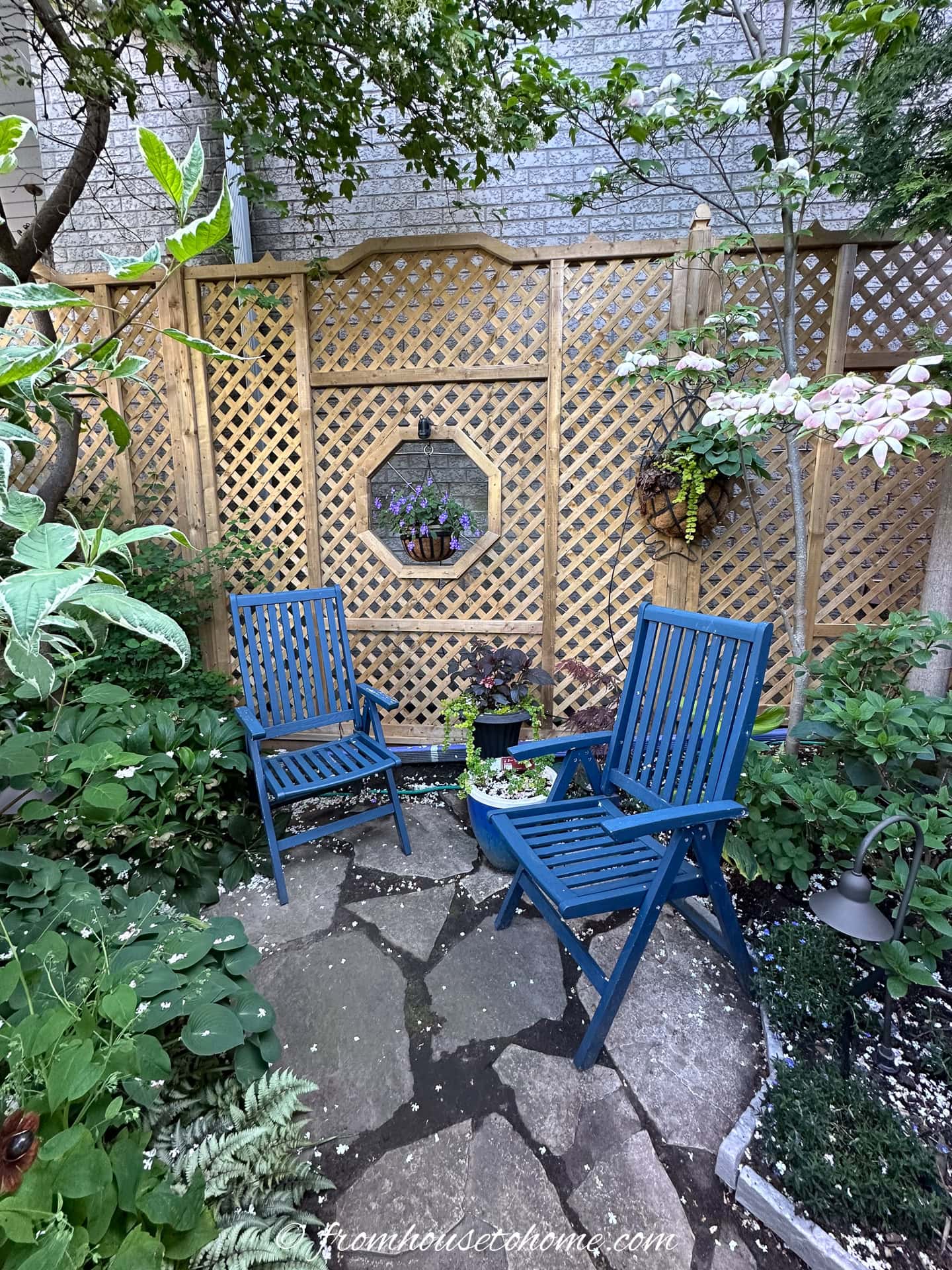 A trellis fence used to block the view of a large brick wall, along with 2 blue garden chairs on a flagstone patio