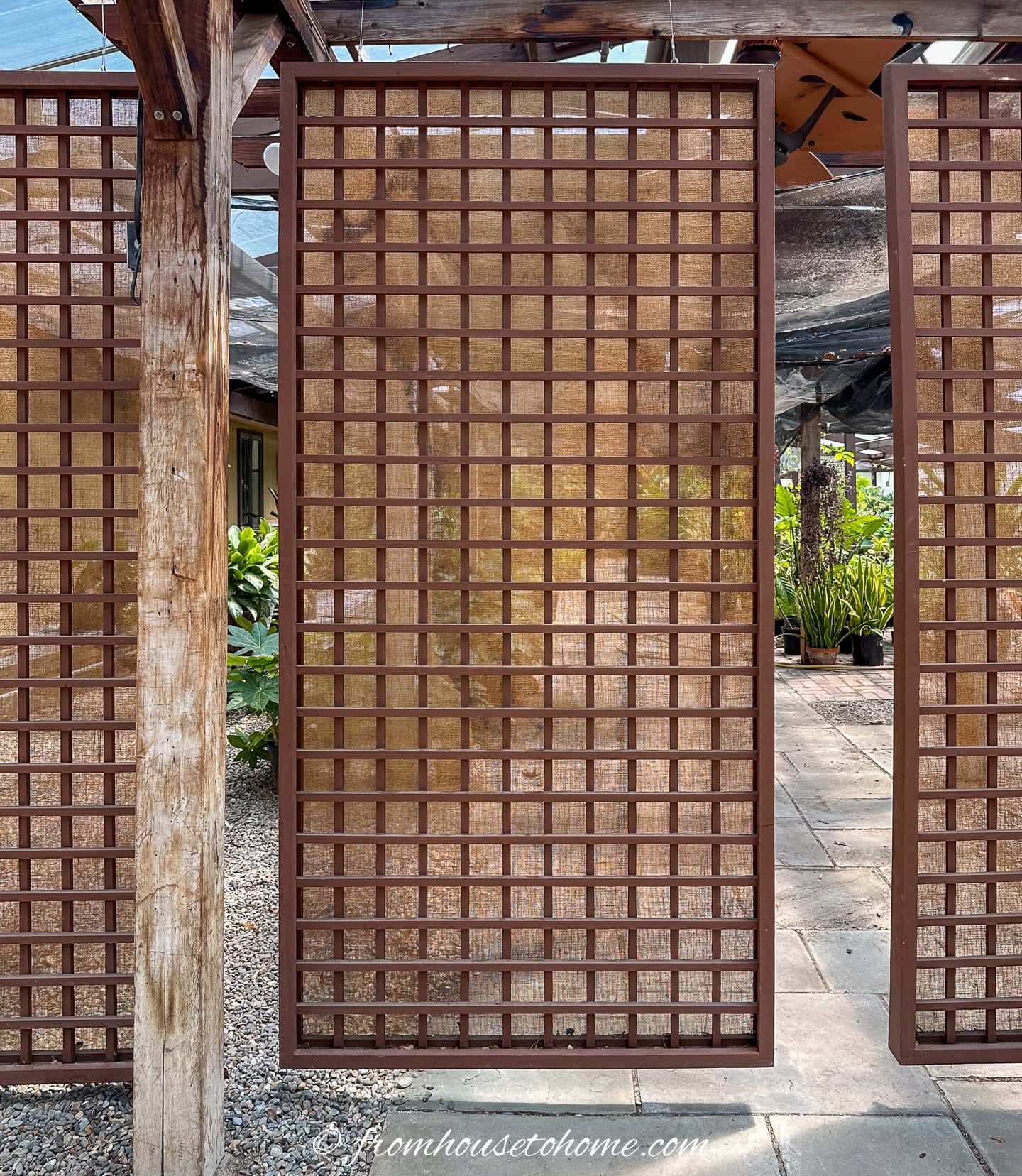 A rectangular wooden trellis panel hangs vertically outdoors, with sunlight filtering through and plants visible in the background.