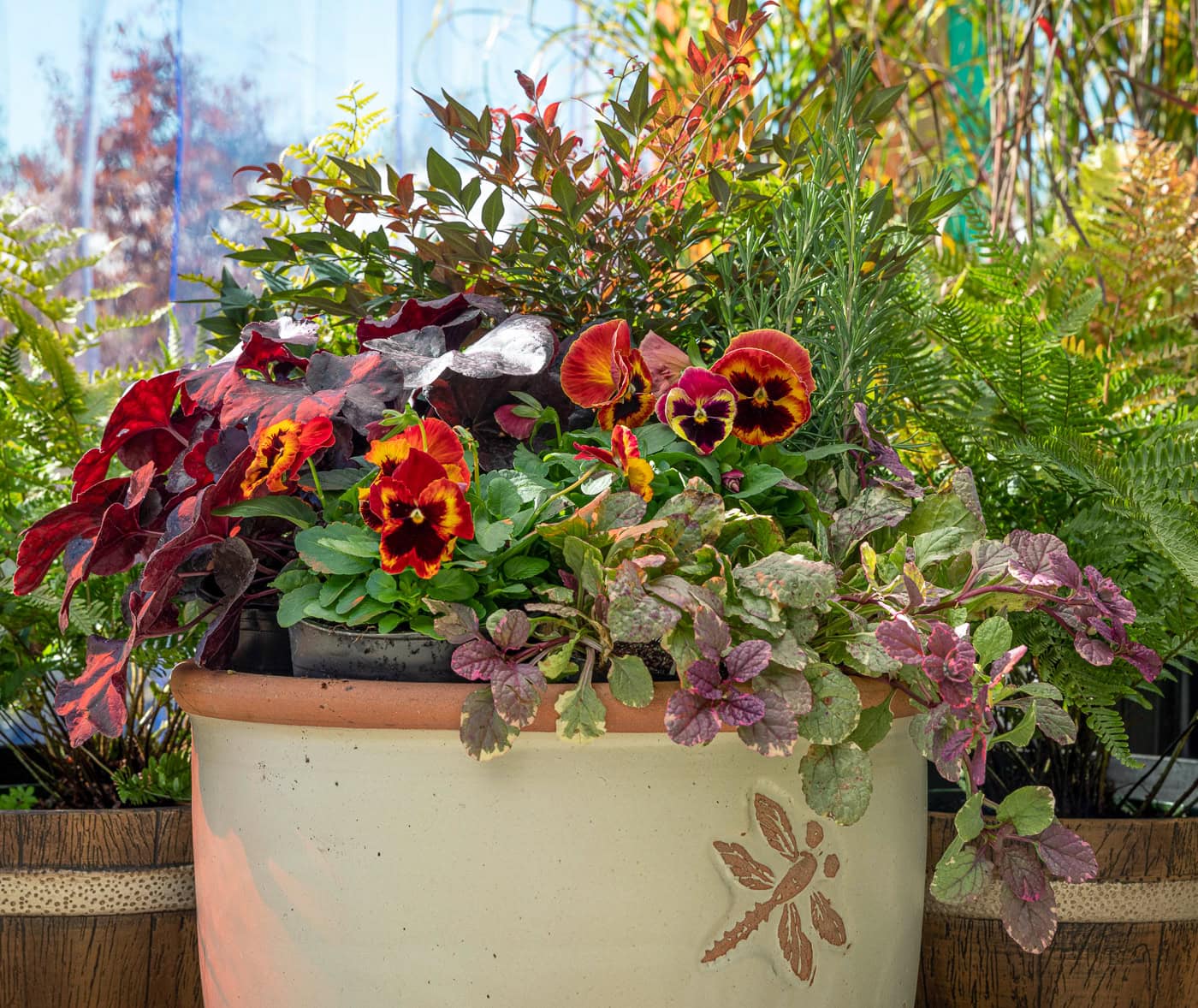 A ceramic planter with a floral design holds a mix of red and yellow pansies, leafy plants, and greenery, set outdoors with sunlight and blurred plants in the background.