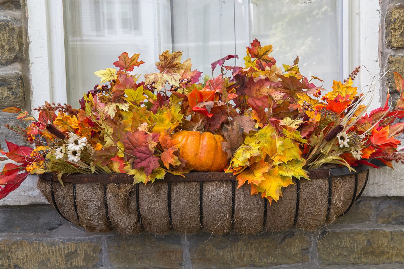 A window box filled with artificial autumn leaves, orange pumpkins, and small decorative flowers set against a stone wall beneath a window.