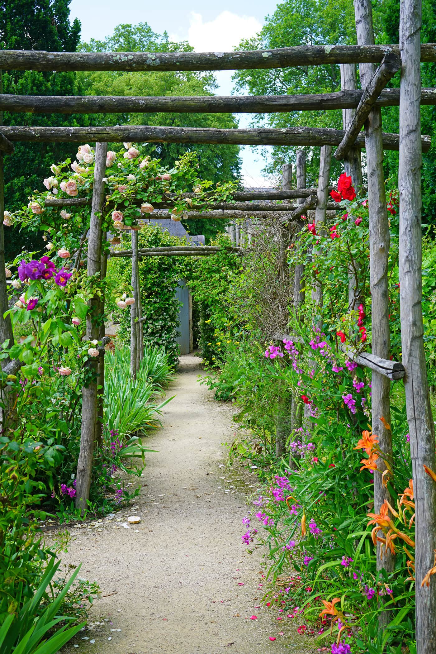 A garden path lined with colorful flowers and greenery, bordered by wooden trellises, leads toward a doorway under a partly cloudy sky.