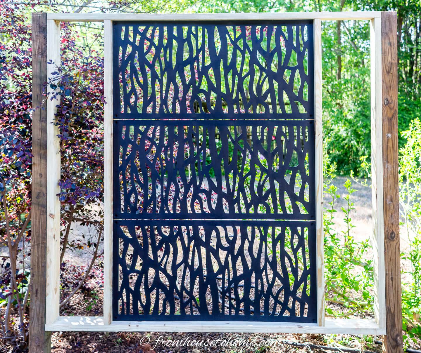Decorative black metal privacy screen with an abstract branch pattern, mounted on a wooden frame outdoors with greenery in the background.