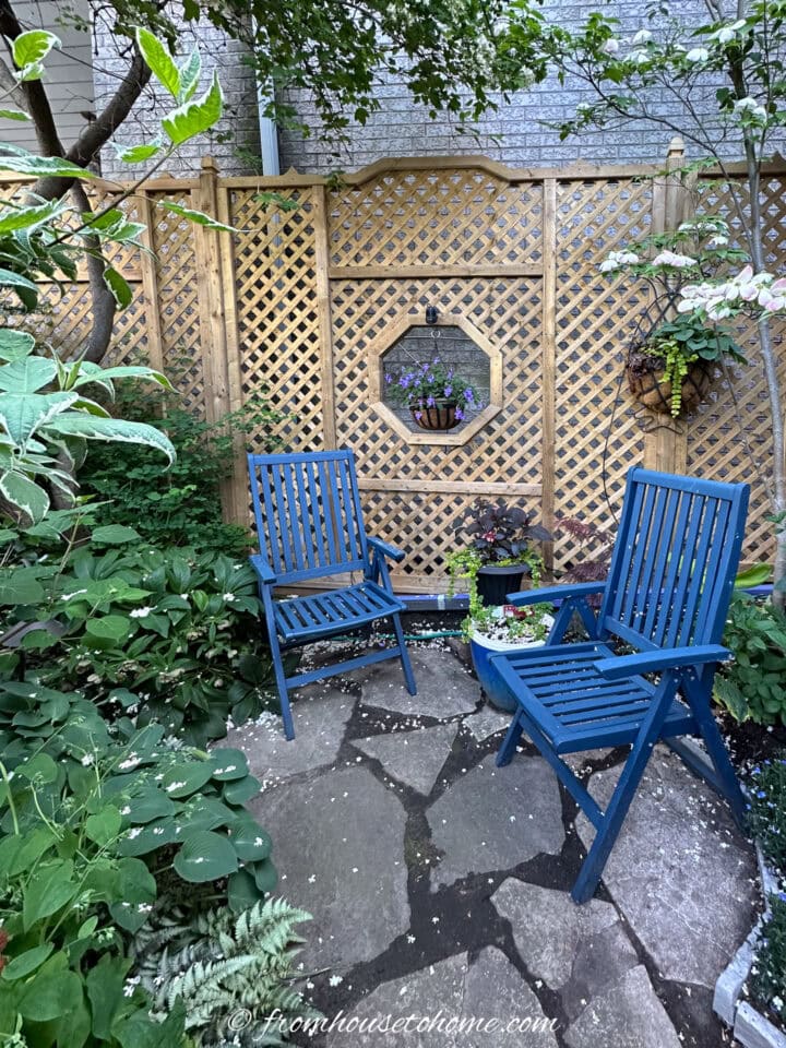 Two blue wooden chairs sit on a stone patio in a small garden, surrounded by green plants and a wooden lattice fence with potted flowers.