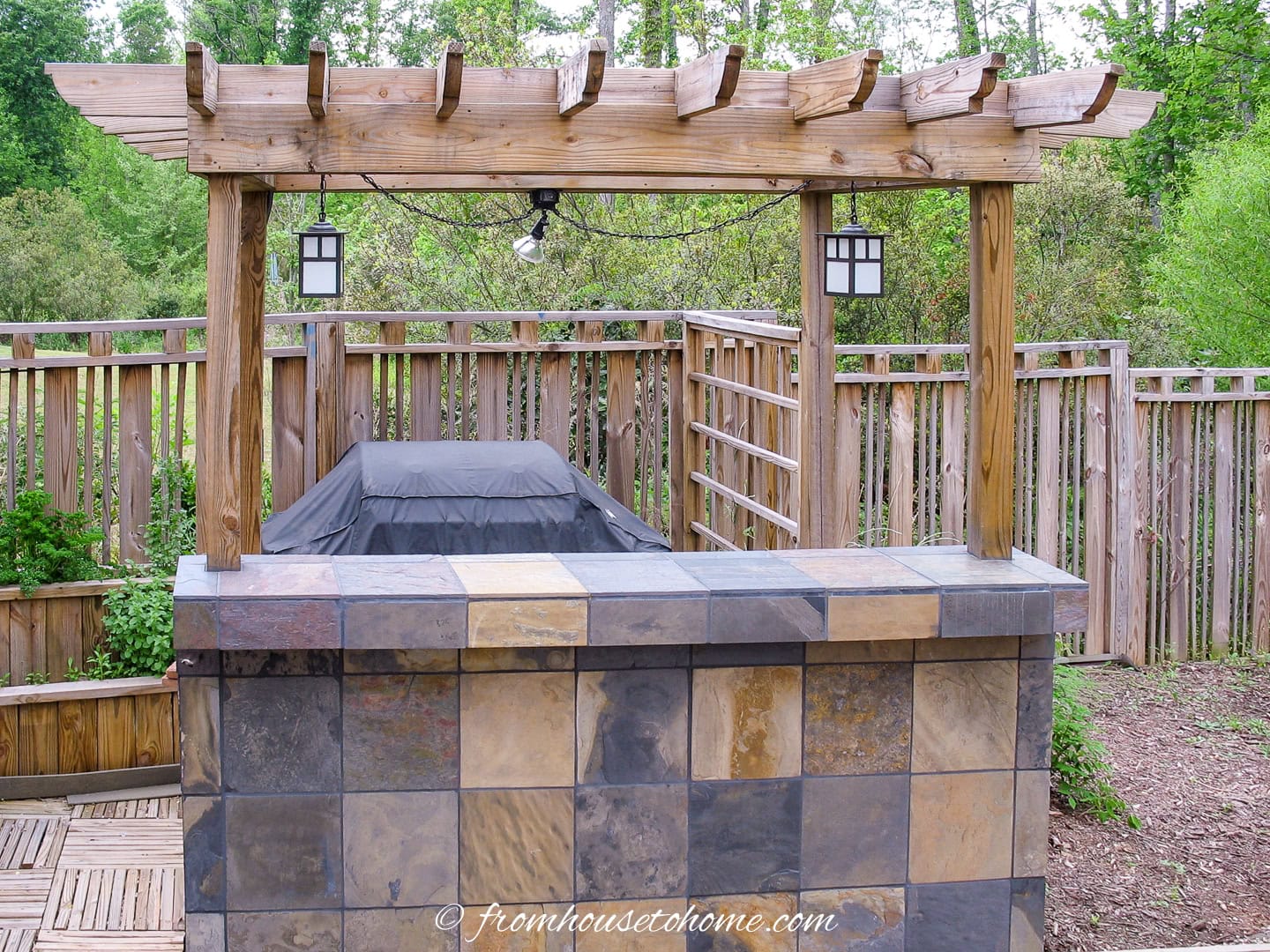 Outdoor grill area with a tiled counter, wooden pergola, hanging lanterns, and a custom privacy fence in the background.
