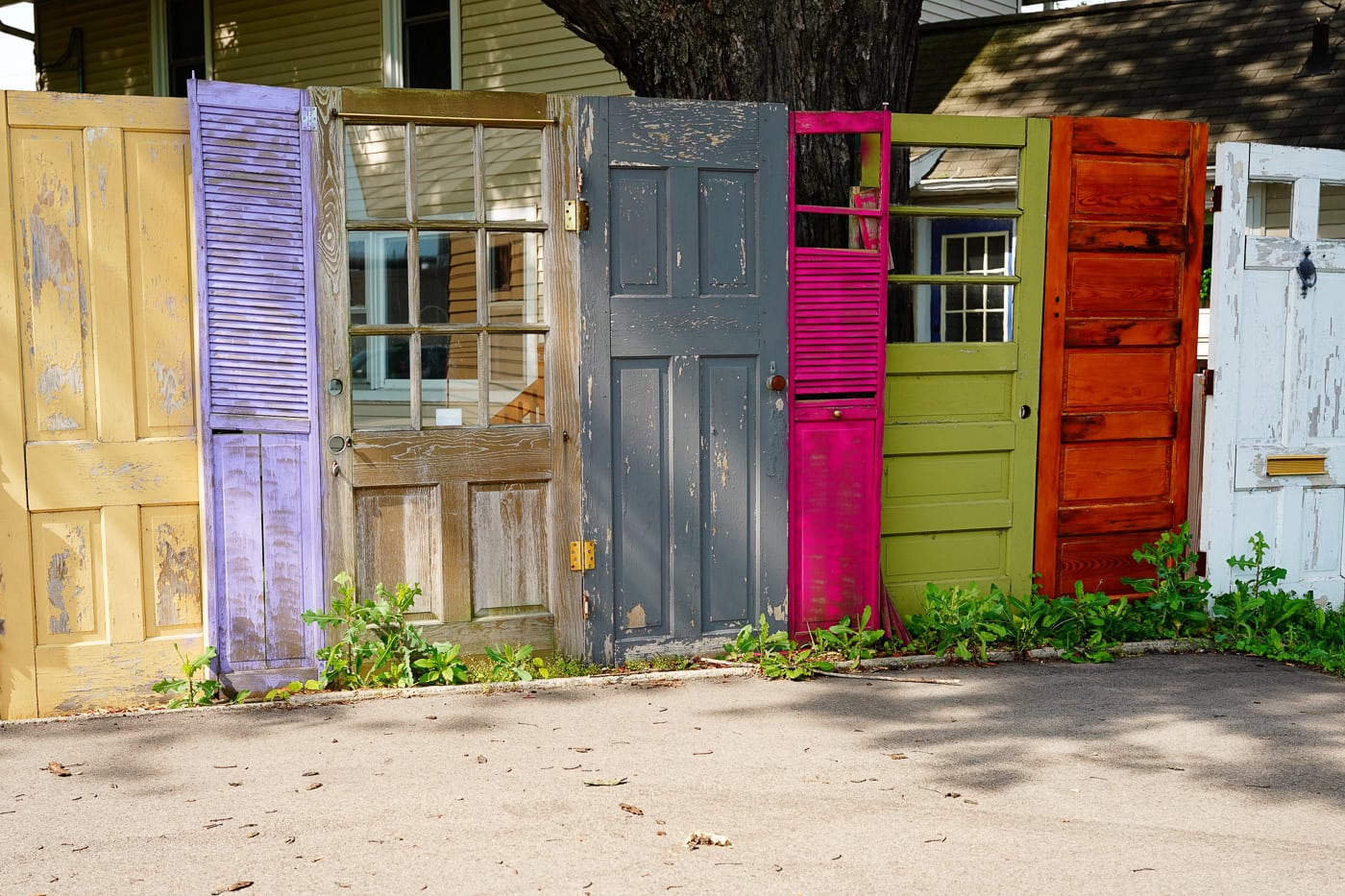 A row of old, colorful doors and shutters is arranged upright outdoors in front of a house, with grass and weeds growing at their bases.