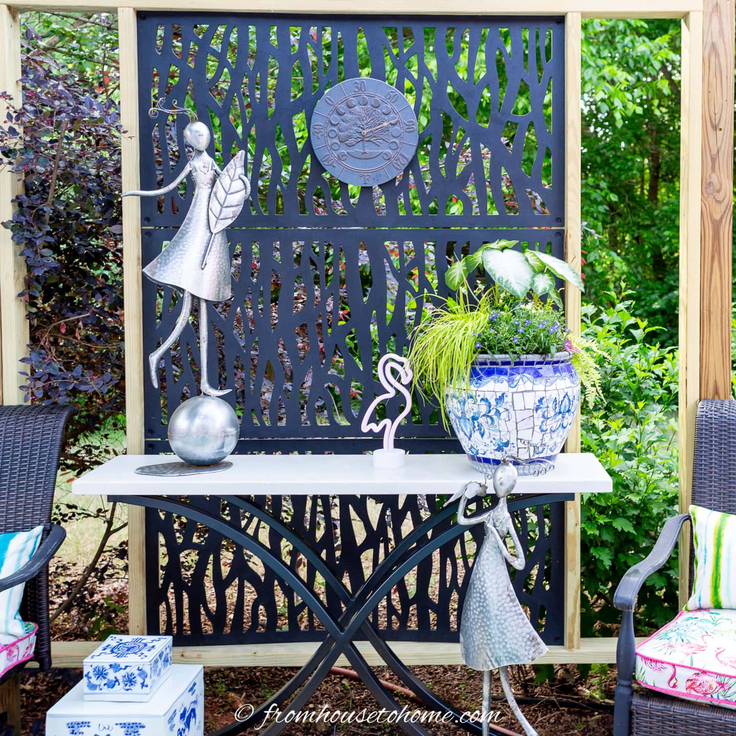 Outdoor garden scene with a decorative black privacy screen, metal fairy sculptures, a white table, a blue and white planter with greenery, wicker chairs, and accent pillows.