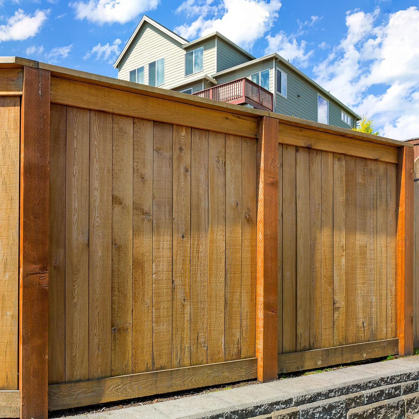 Wooden privacy fence with vertical planks and sturdy posts in front of a modern two-story house under a partly cloudy sky.