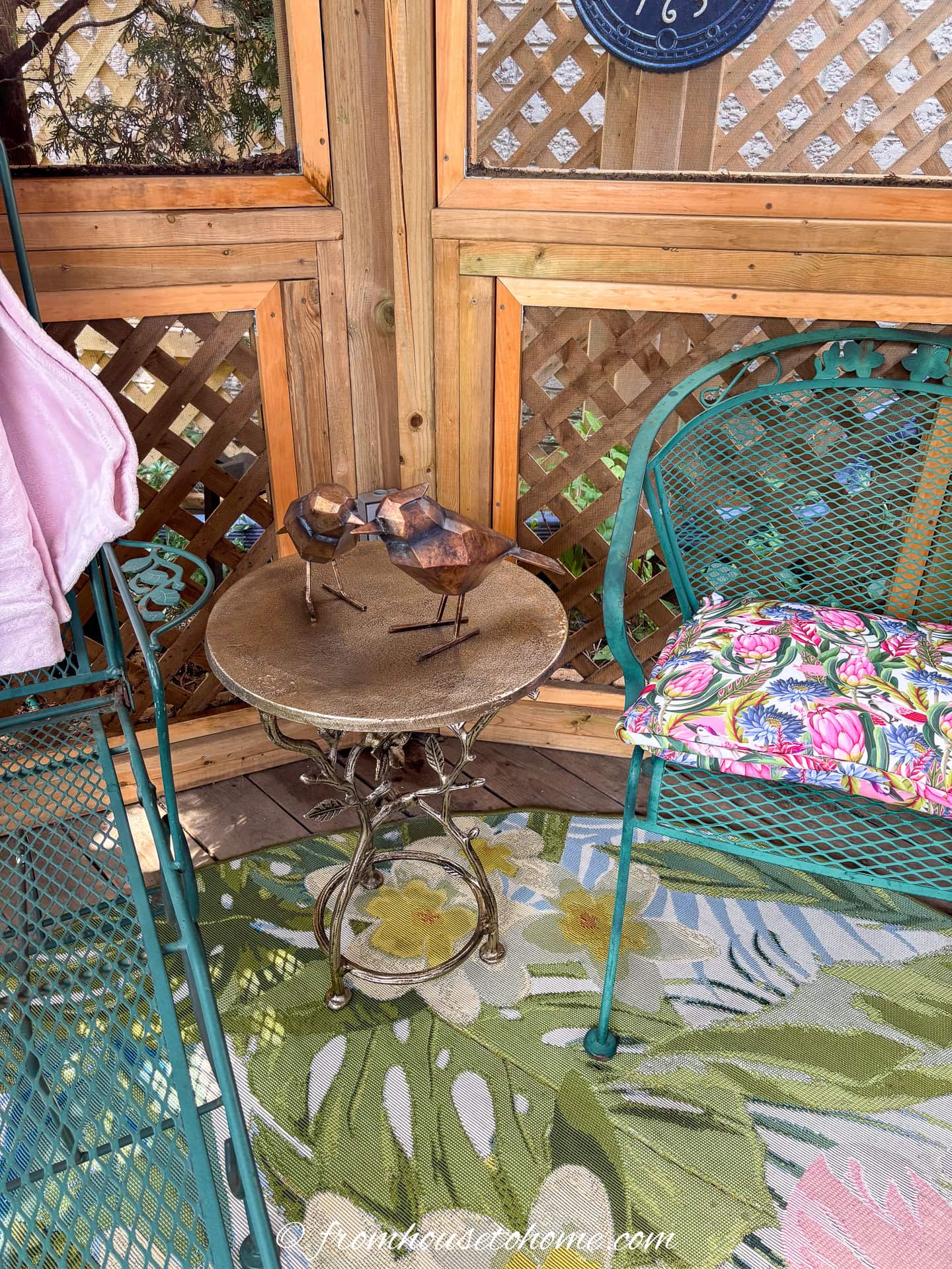 A small round table with bird sculptures stands between two green metal chairs with floral cushions on a patterned rug in a wooden gazebo.