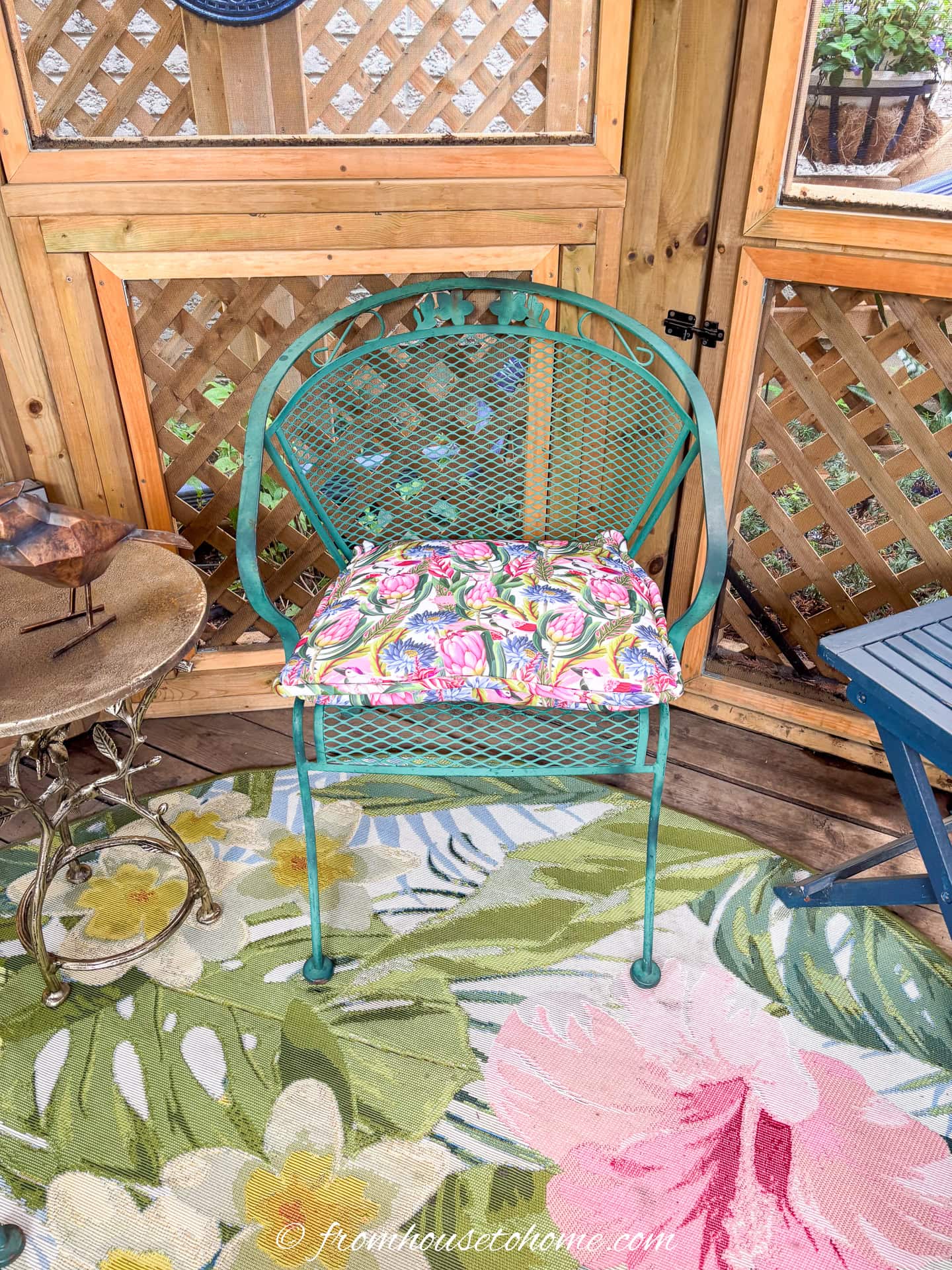 A green wrought-iron chair with a pink and blue cushion on a pink, blue and green outdoor rug in a gazebo