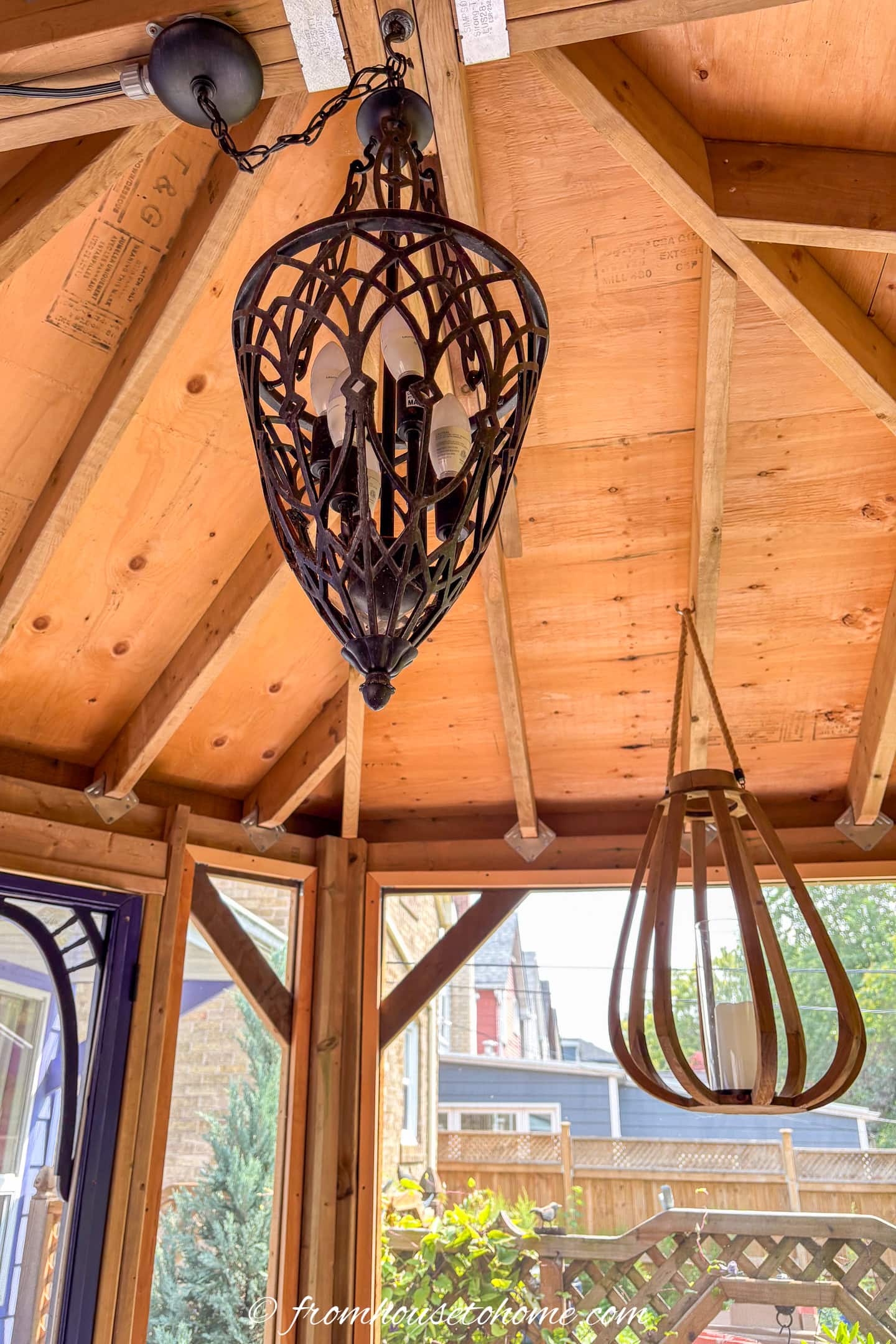 Two pendant light fixtures hang from the wooden ceiling of a sunroom with visible rafters and a view of houses and greenery outside.