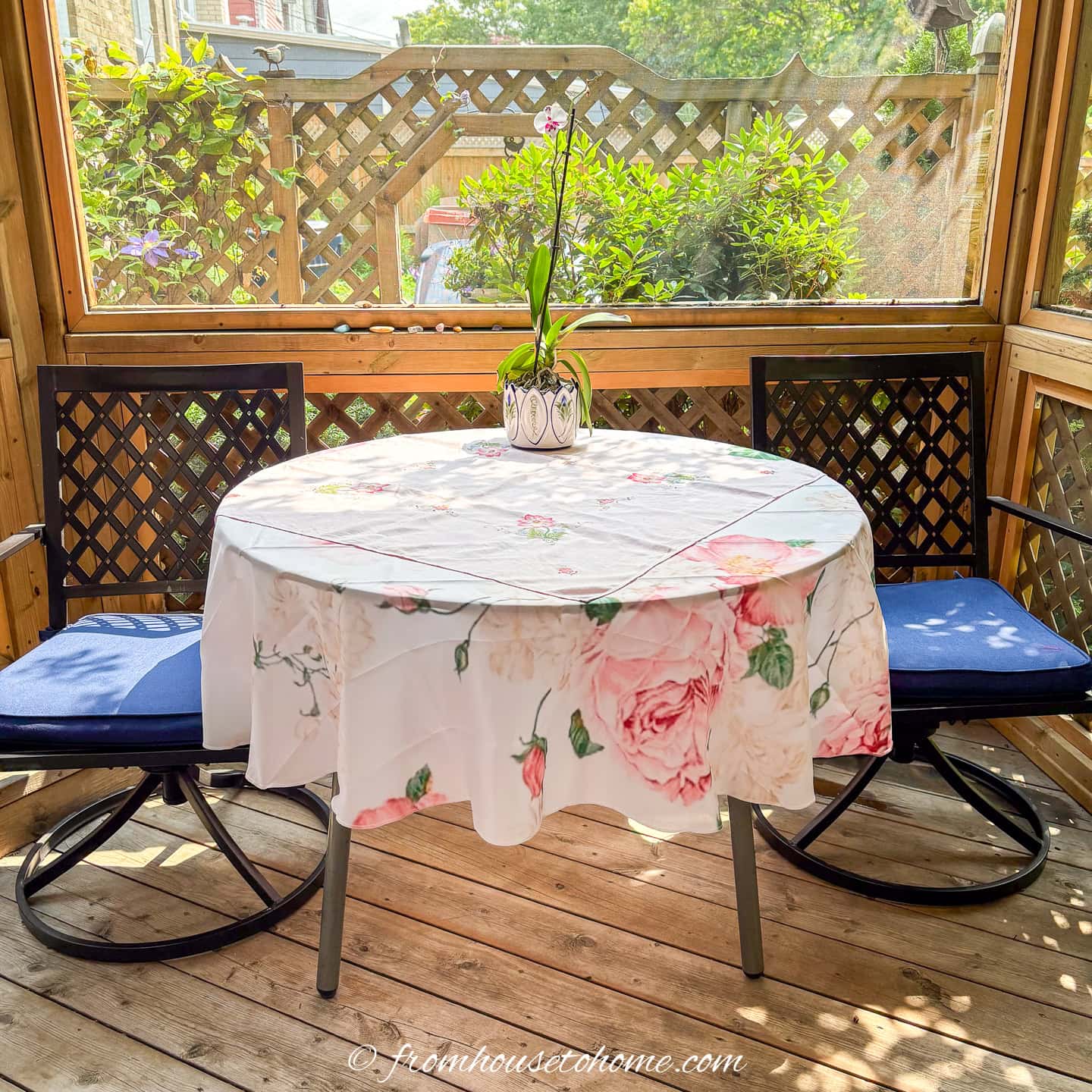 A small round table with a floral tablecloth and a potted orchid is set between two blue cushioned chairs on a wooden patio.
