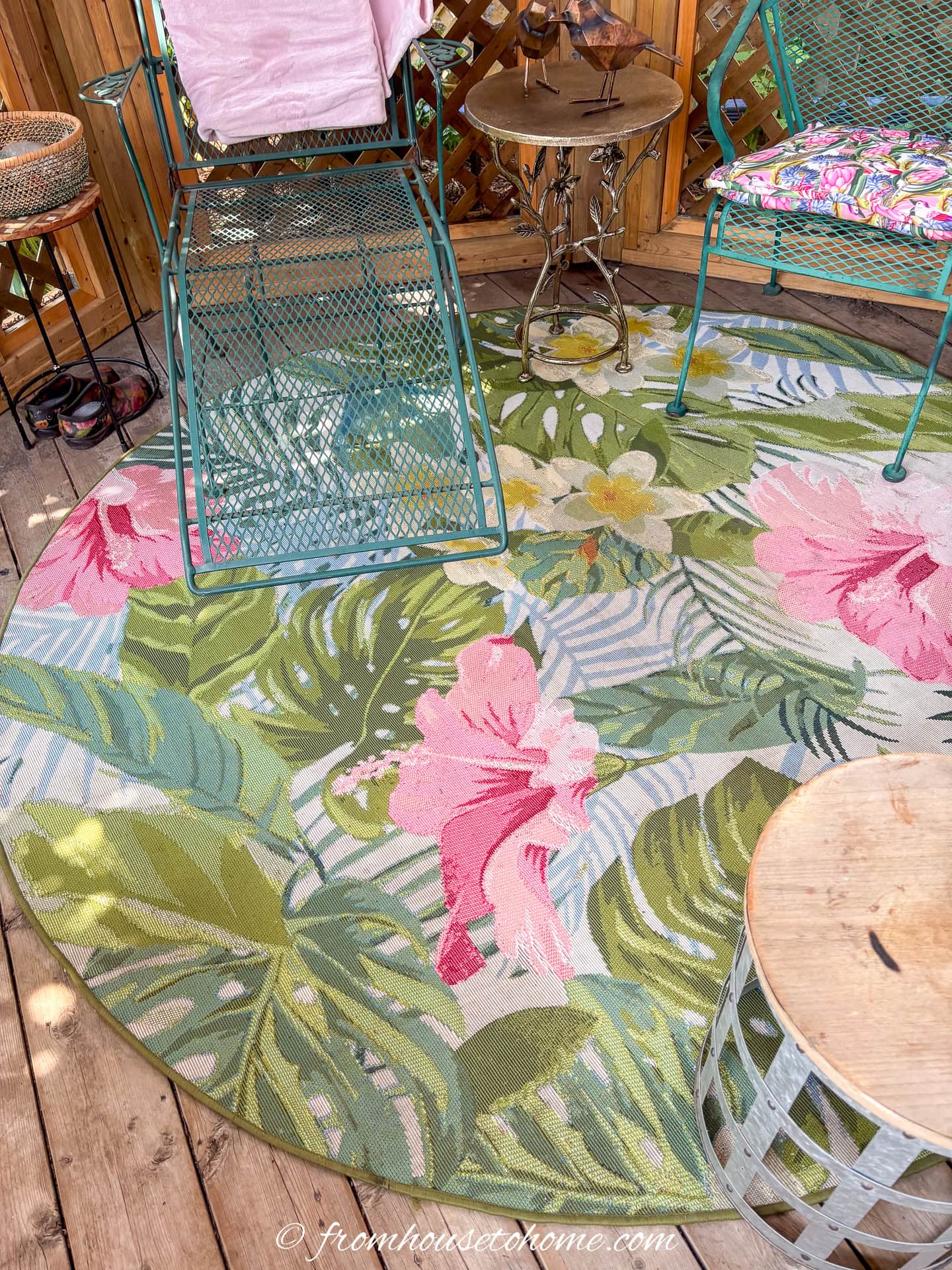 Round outdoor rug with a pink and green tropical floral pattern, surrounded by green metal patio furniture and a wooden deck.