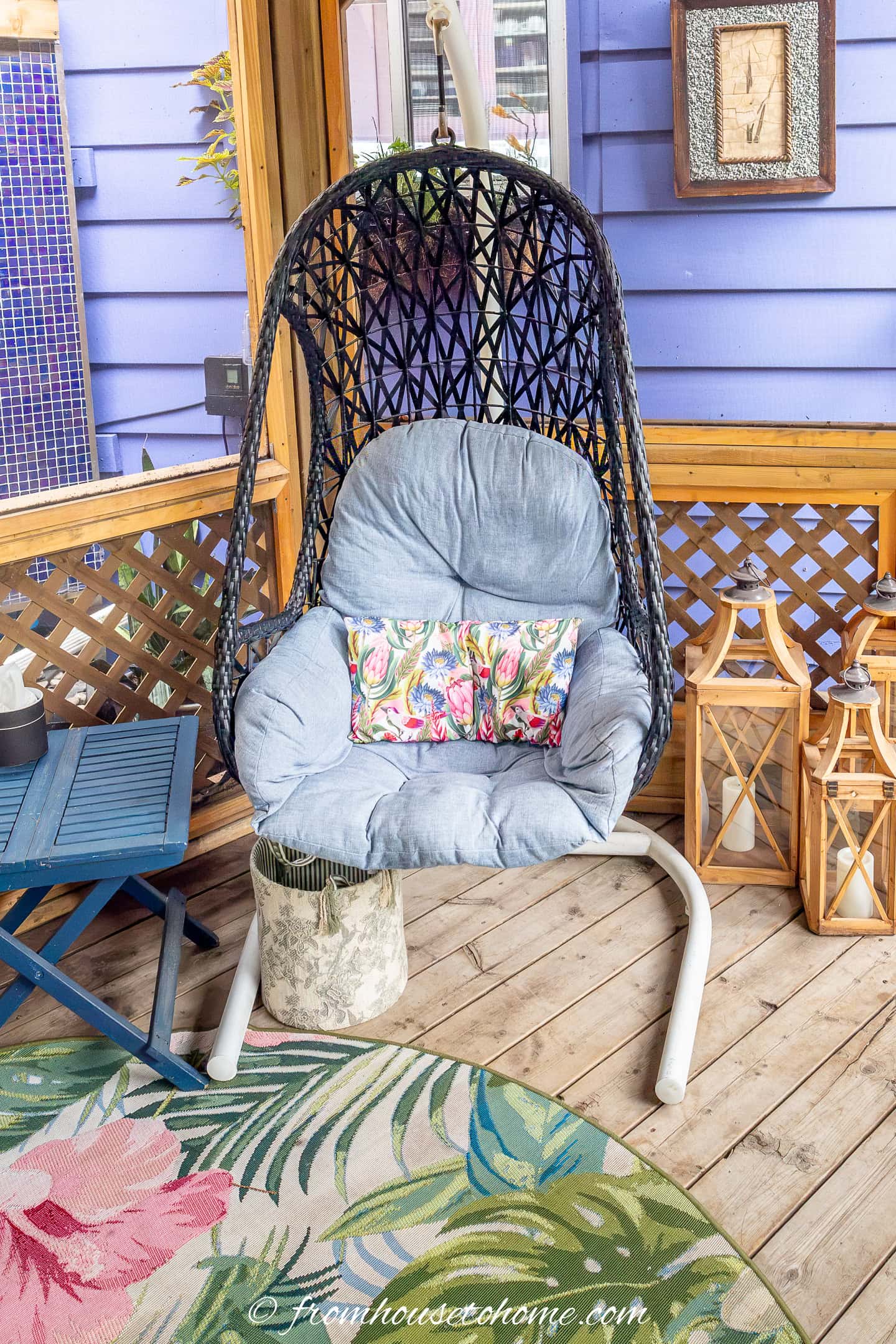 Cushioned hanging chair with a floral pillow on a wooden porch, next to lanterns, a blue table, and a round floral rug.