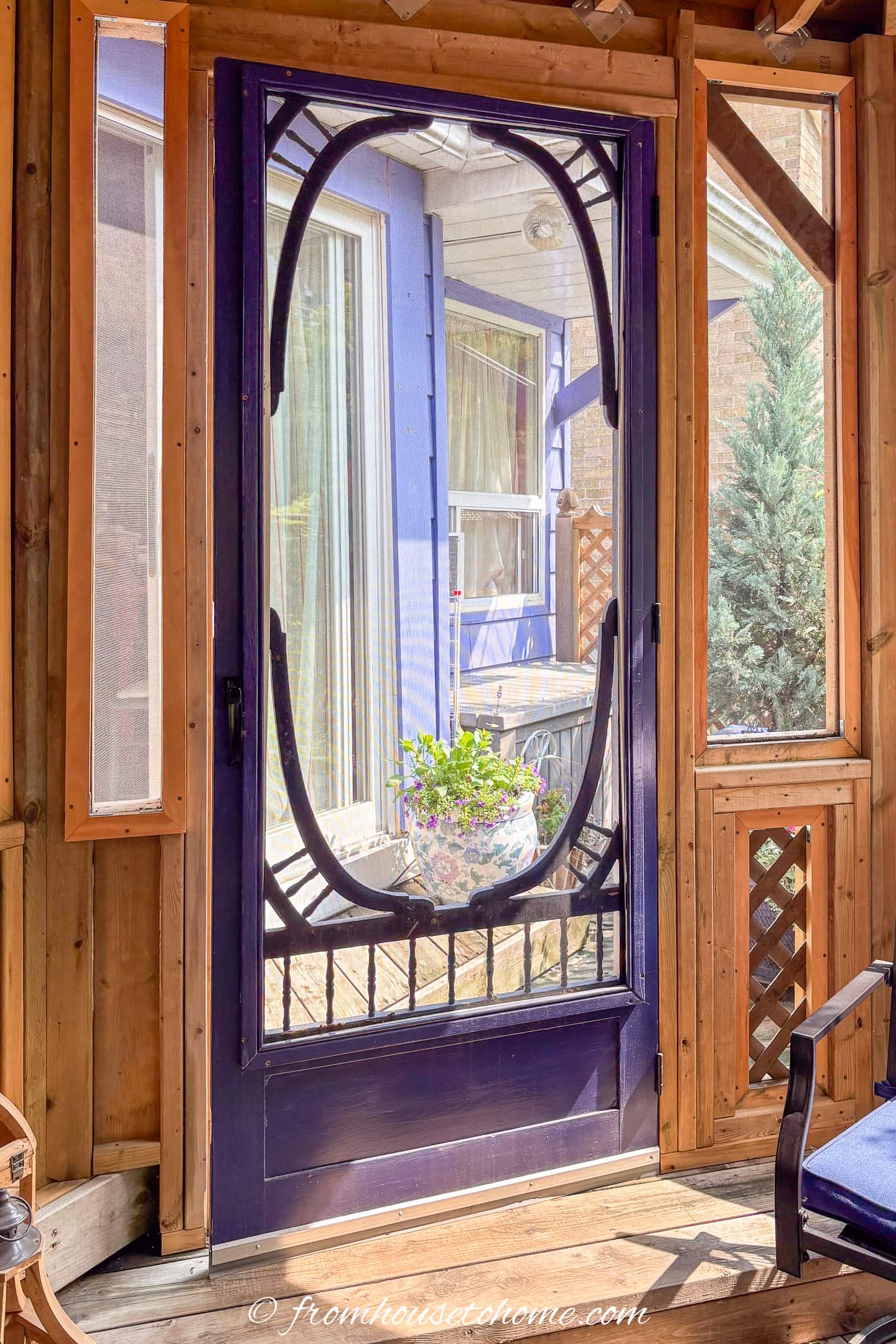 A purple screen door with decorative trim opens to a view of a blue house, a window with yellow curtains, and a potted plant on the porch.