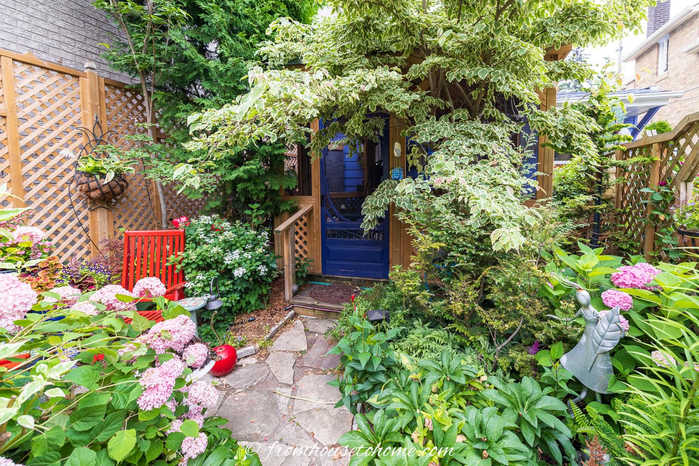 Small garden with stone path, lush plants, pink hydrangeas, lattice fencing, red chair, and a wooden shed partially hidden by greenery.