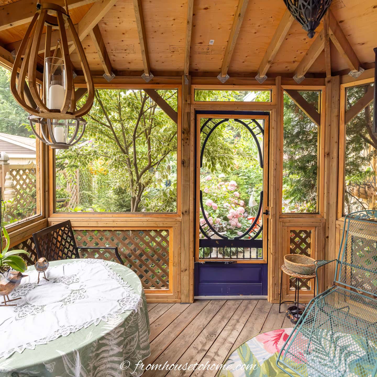 A screened porch with a wooden ceiling, a round table covered by a tablecloth, wire patio chairs, potted plants, and a door opening to a garden with trees and flowers outside.