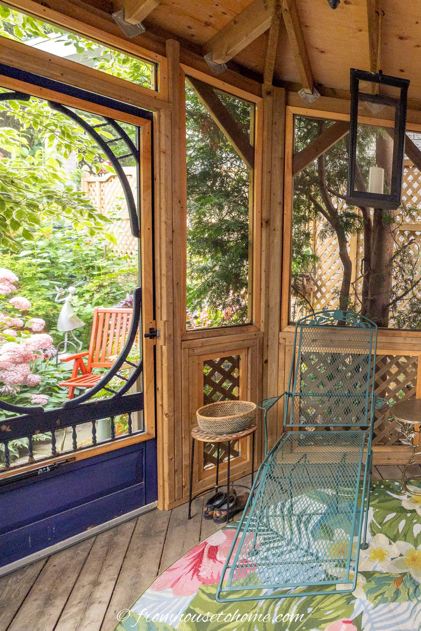 Screened-in porch with a metal lounge chair, small side table, and floral rug, overlooking a lush garden with flowers, a red bench, and garden decor through a circular window.