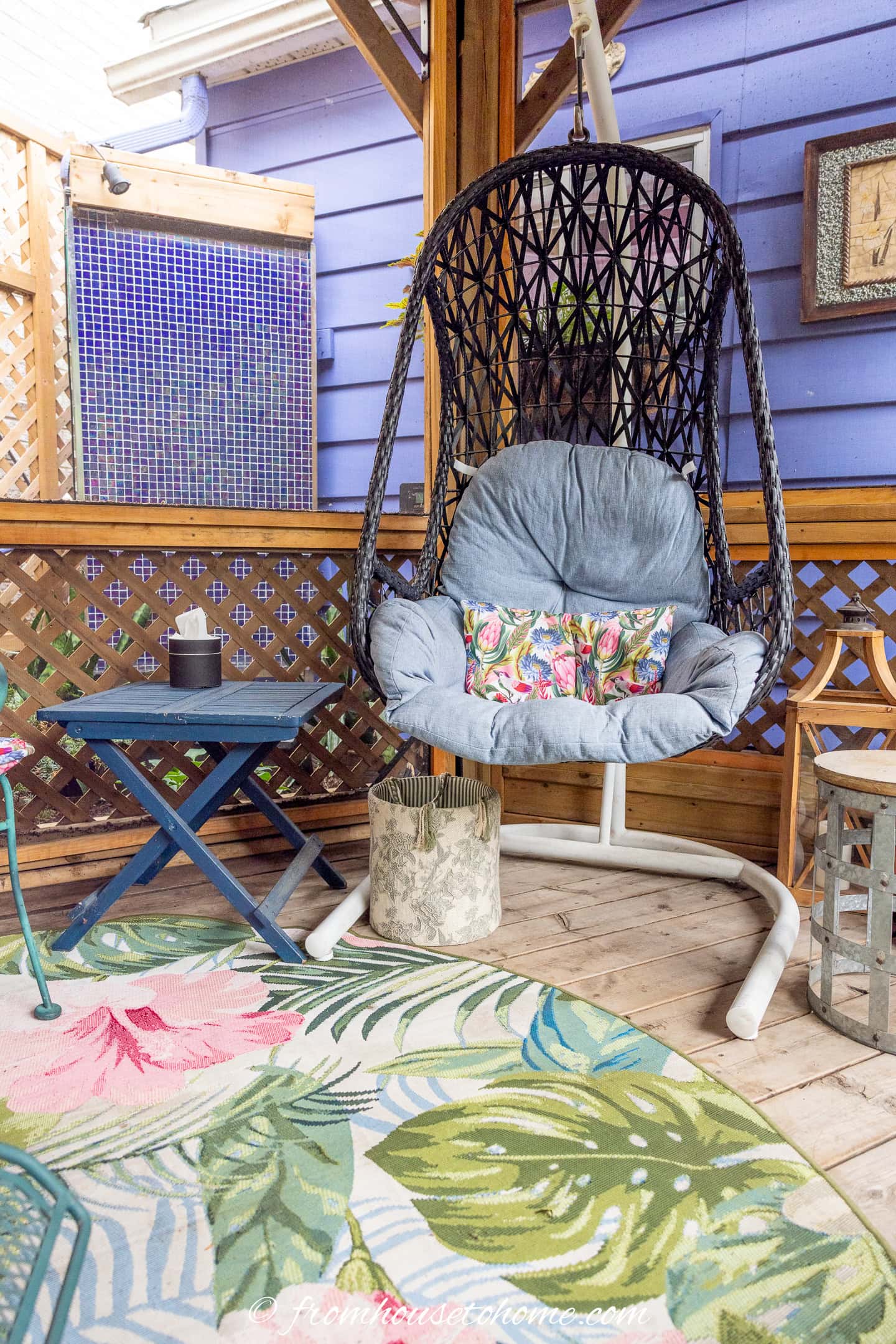Wicker hanging chair with blue cushion and floral pillow on a wooden patio, next to a blue table and tropical-patterned rug.