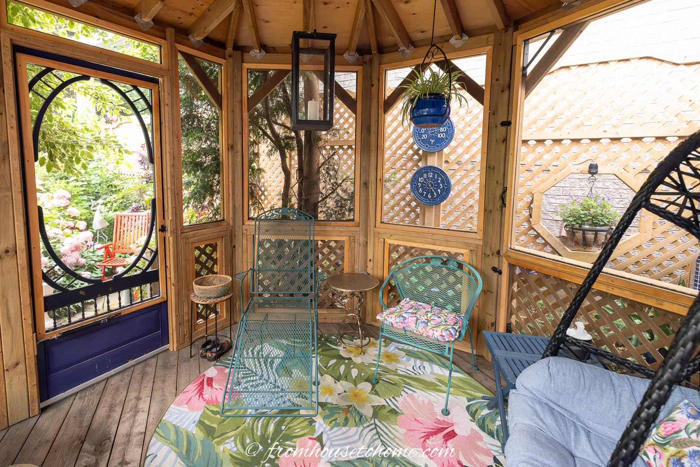 Wooden gazebo interior with green metal chairs, floral rug, hanging plants, decorative plates, and lattice walls. An open door reveals greenery and a red chair outside.