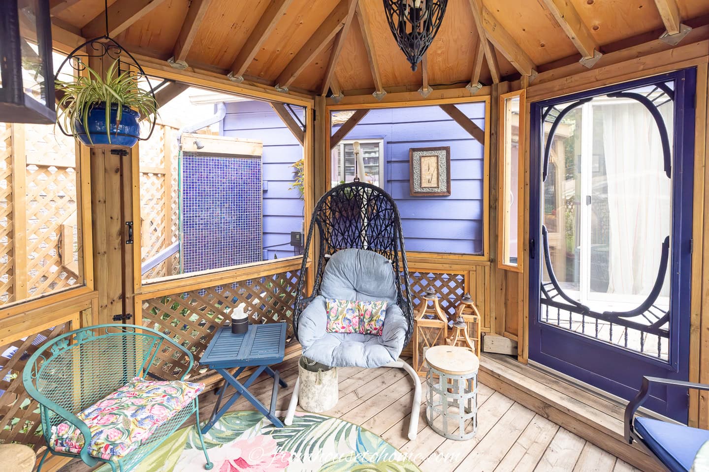 Wooden gazebo with a hanging chair, small tables, potted plants, and a view of a purple house through lattice walls and windows.