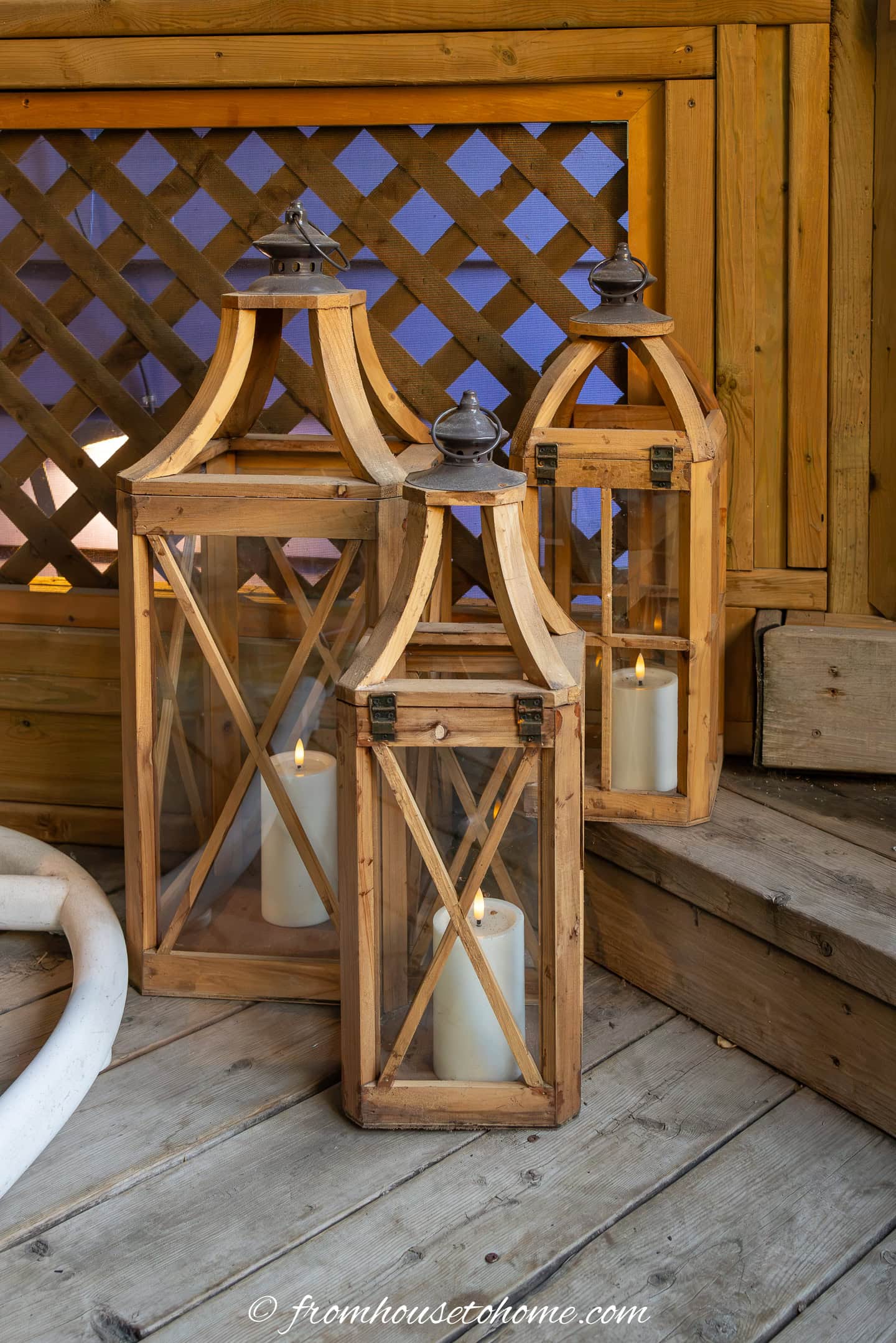 Three wooden and glass lanterns with lit candles inside are placed on a wooden deck next to steps and a lattice fence.