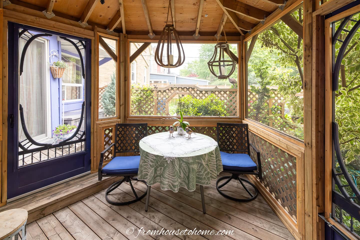 Wooden gazebo with a round table covered by a lace cloth, two blue-cushioned chairs, hanging lights, and a view of a garden through screened windows.