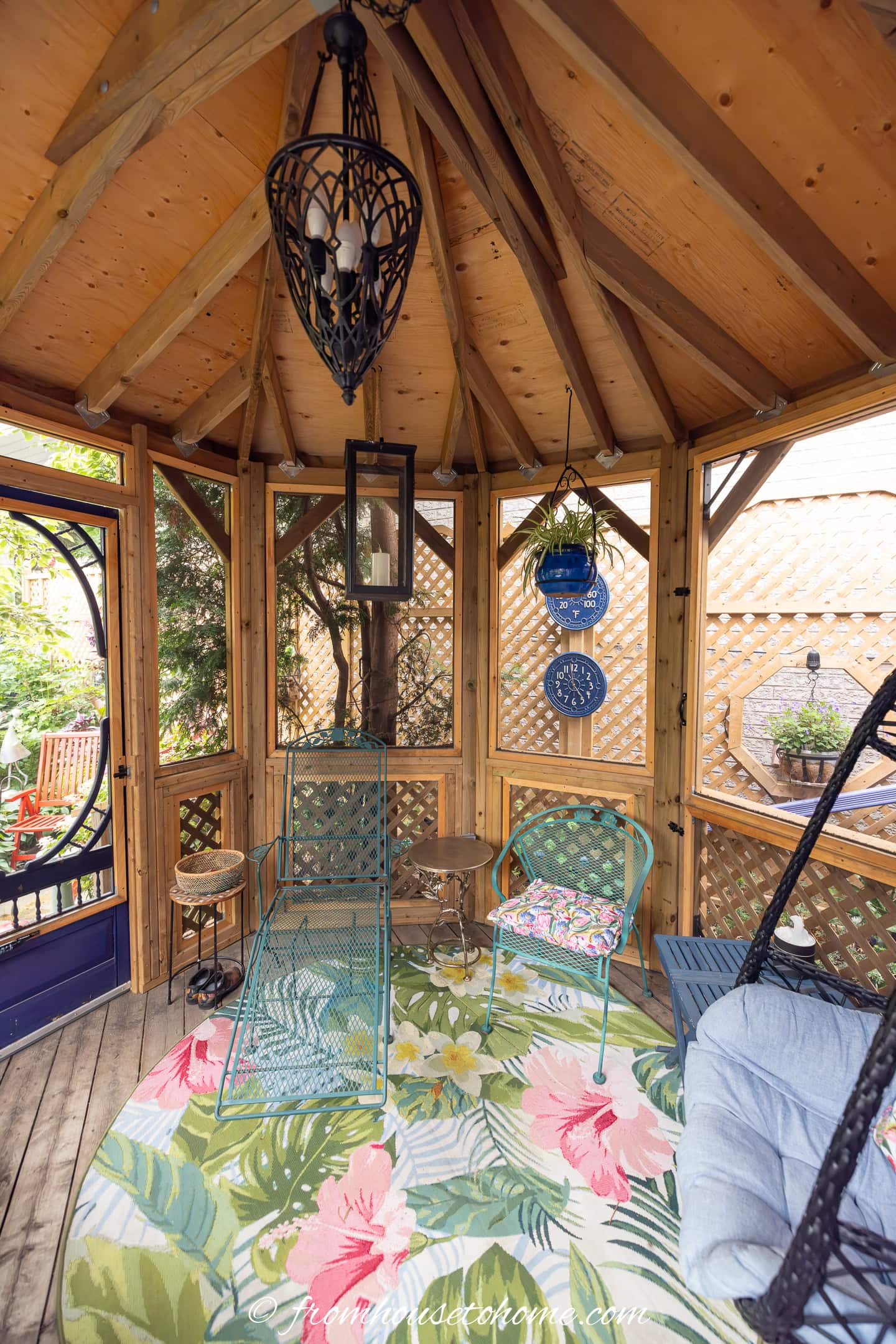 Screened wooden gazebo with a vaulted ceiling, metal chairs, a floral rug, hanging blue plates, and garden views through lattice walls.