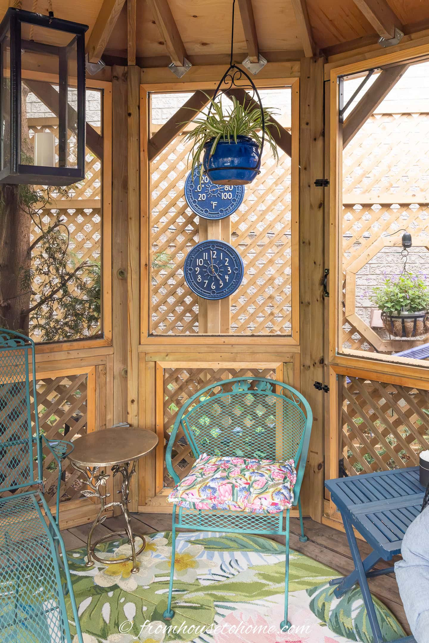 A wooden gazebo with lattice walls, a hanging blue planter, blue metal chair with a floral cushion, a small table, and two wall-mounted weather dials showing temperature and humidity.