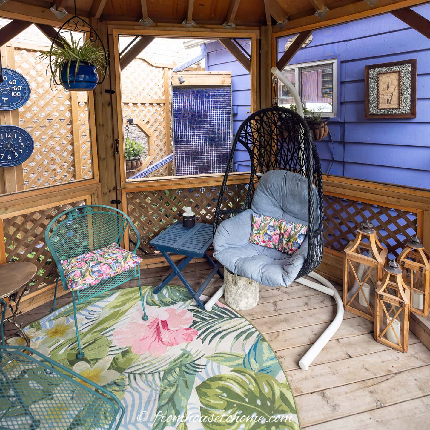 A small wrought iron chair beside a hanging basket chair and some lanterns in a gazebo with an outdoor rug