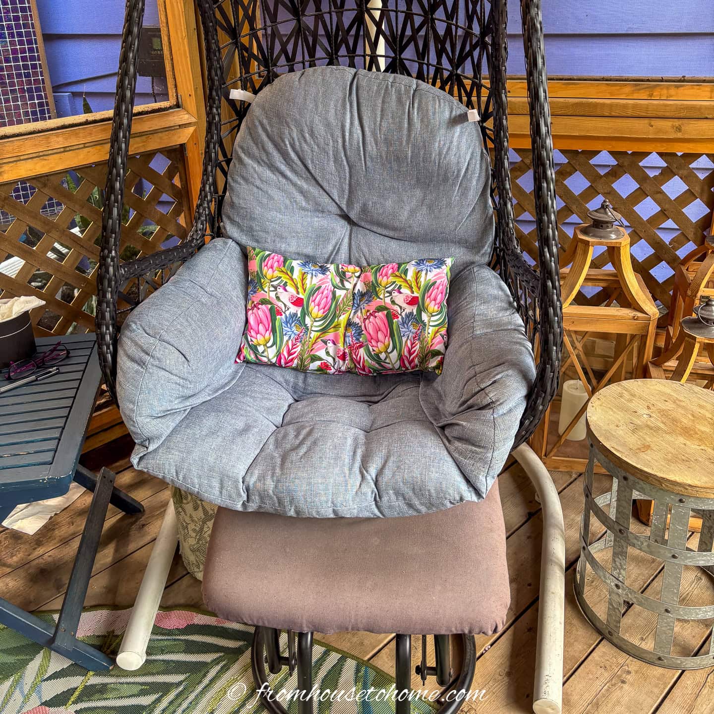 Gray cushioned hanging chair with a colorful floral pillow on a wooden porch, surrounded by small tables and lattice walls.