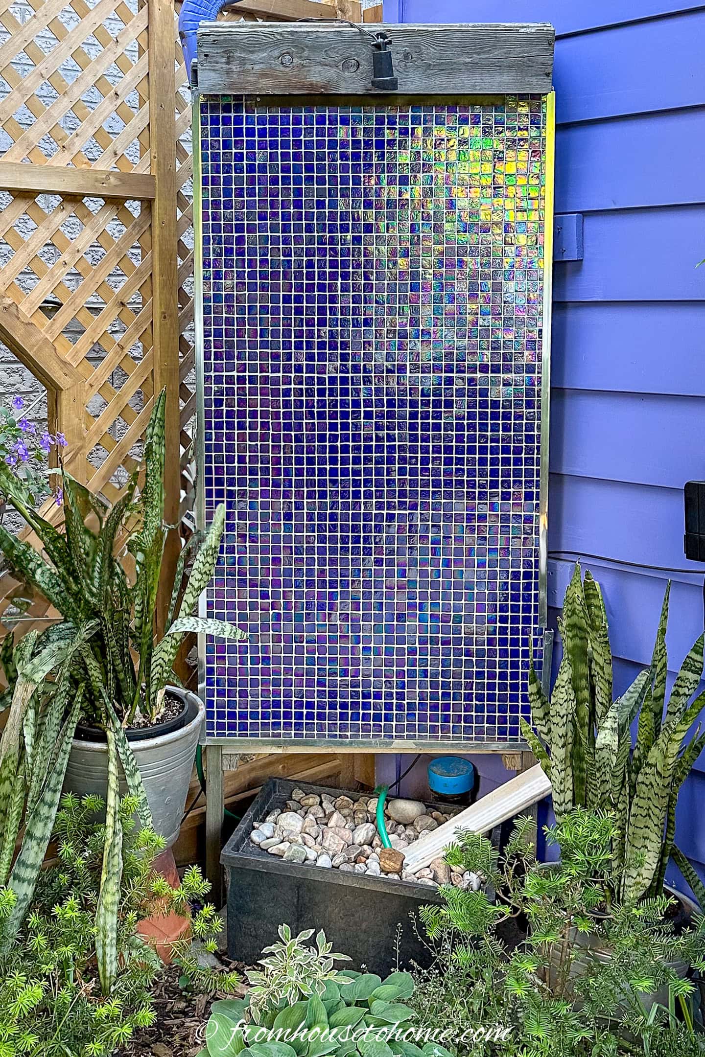 A rectangular mosaic of small blue tiles stands against a purple wall, surrounded by potted plants and a container of rocks and water below.
