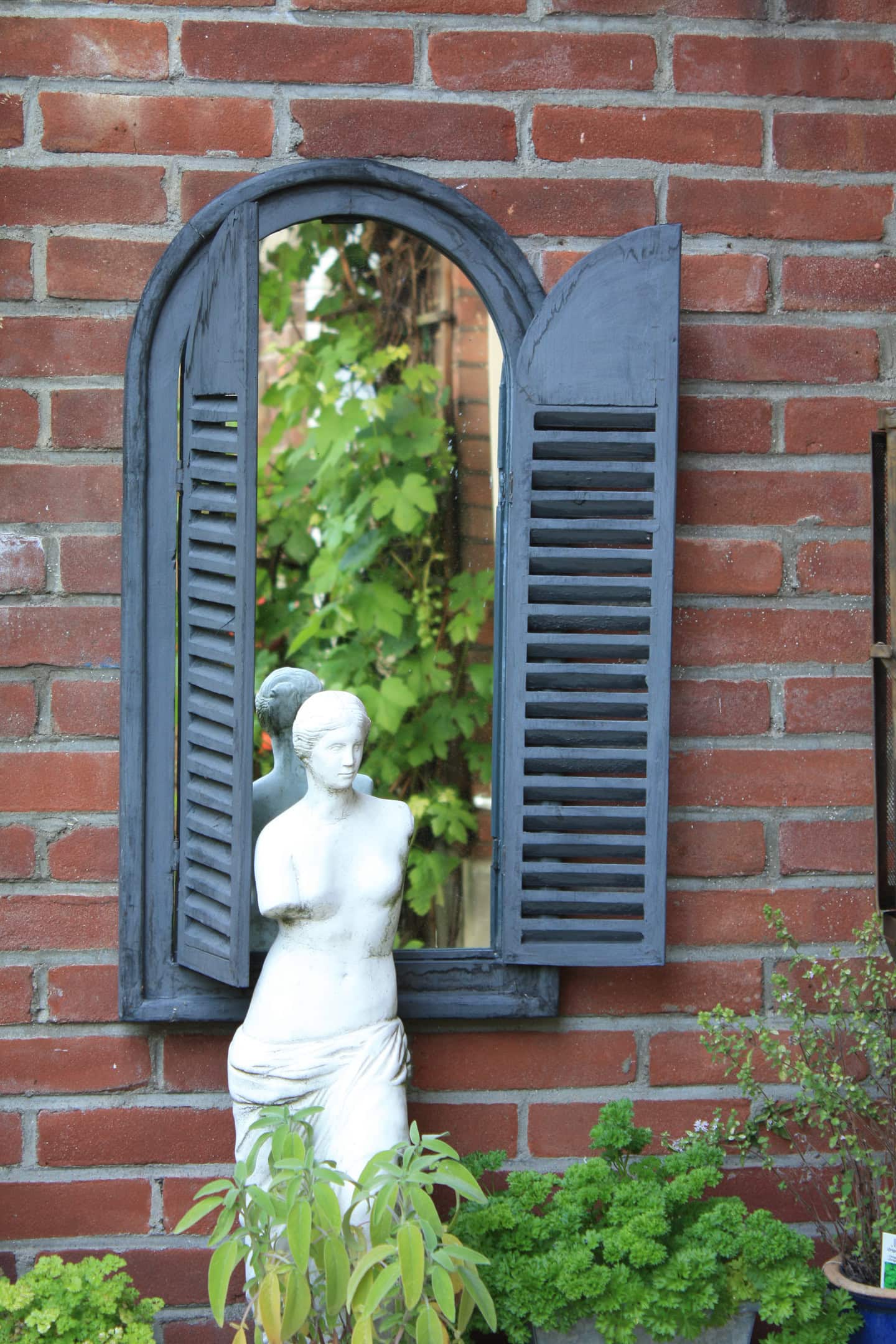 A white statue of a woman stands in front of a brick wall with a window featuring black shutters and a mirror reflecting green foliage.