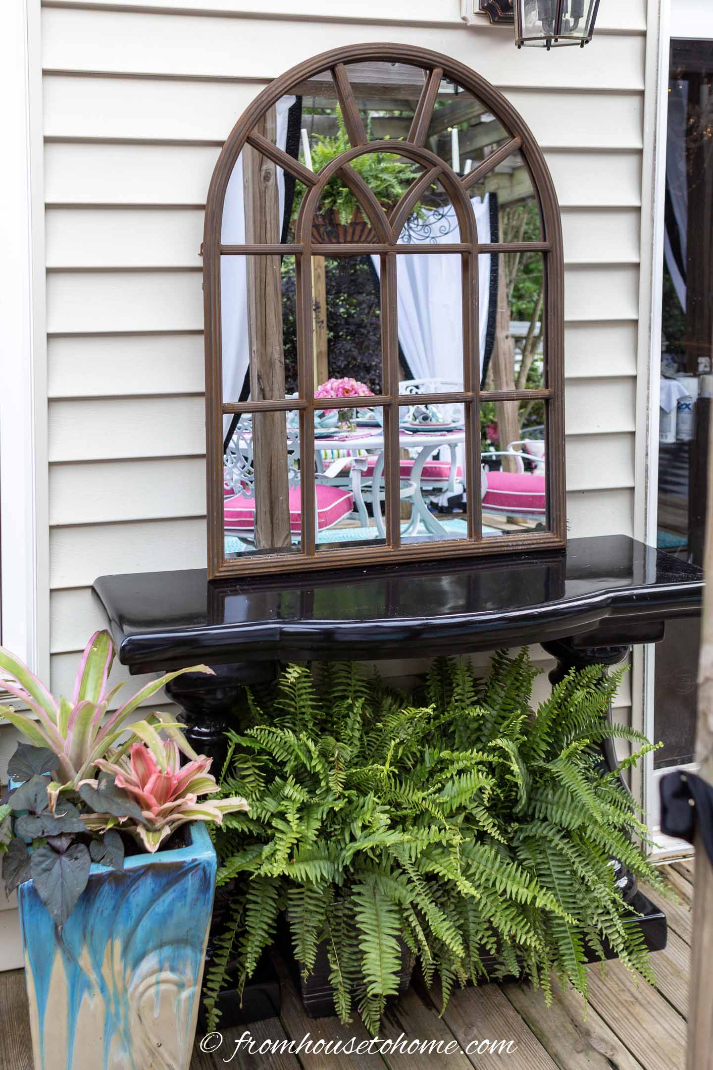 A large arched mirror sits on a black table with a fern underneath; a blue planter with tropical plants is on the left; reflections of outdoor furniture appear in the mirror.