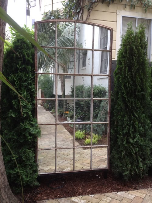 A large, multi-pane mirror stands outdoors between two tall bushes, reflecting a garden path, plants, and part of a house exterior.