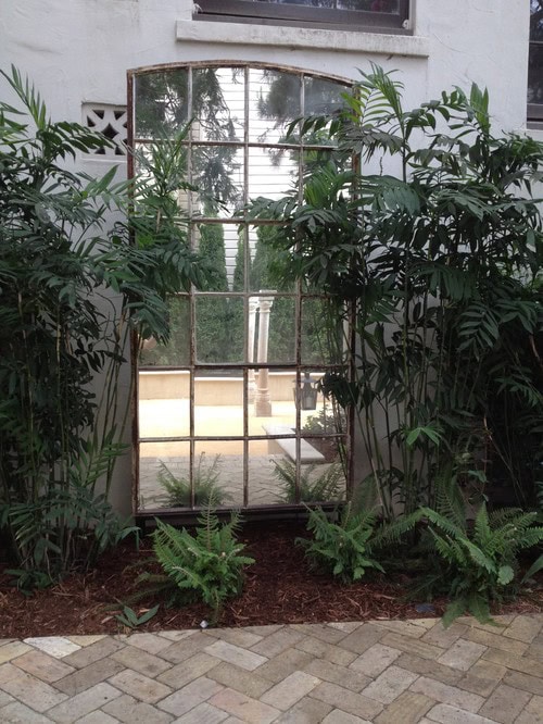 A large, tall mirror framed like a window is mounted on an exterior wall, surrounded by green plants and ferns.