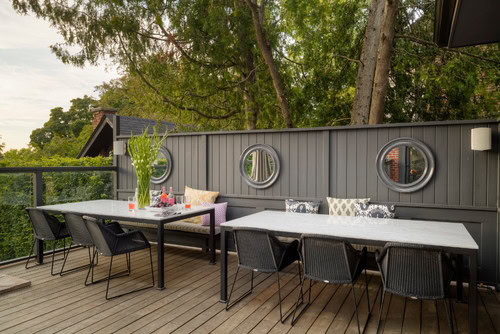 Outdoor patio with two tables, black chairs, gray wooden fence with round mirrors, cushions, and green plants, surrounded by trees.