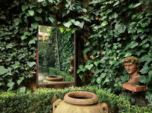 Outdoor garden scene with dense ivy, a wall-mounted mirror reflecting greenery, a terracotta urn in front, and a classical-style bust on a brick ledge to the right.
