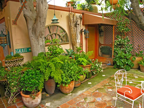 A cozy patio with potted plants, tiled flooring, a white metal chair with a red cushion, and various wall decorations near a wooden door.