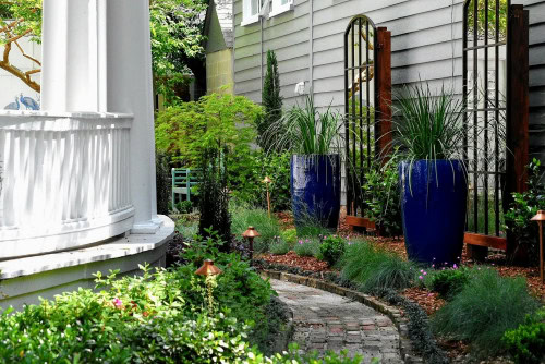 Curved brick path bordered by plants, large blue planters, and trellises in a landscaped side yard beside a gray house and white porch.
