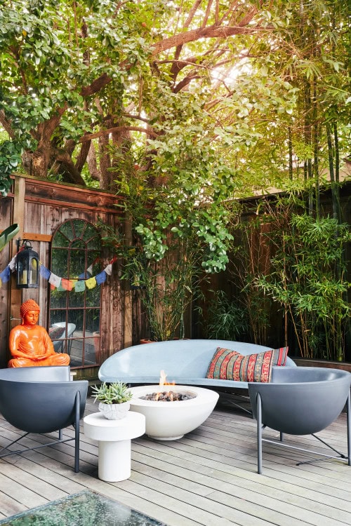 Outdoor patio with modern chairs, a round fire pit, a curved sofa with a colorful pillow, a potted plant, an orange Buddha statue, prayer flags, and trees providing shade.