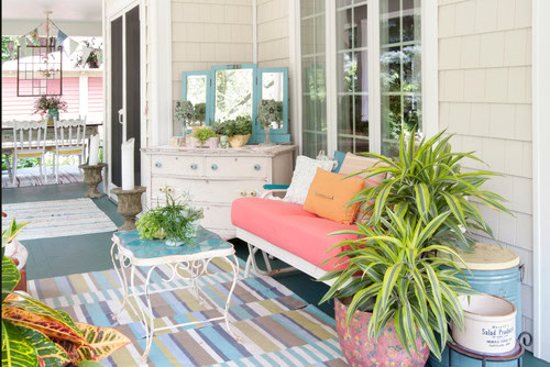 A porch with a white bench and coral cushion, striped rug, potted plants, small table, and a white dresser with mirrors, set against beige siding and large windows.