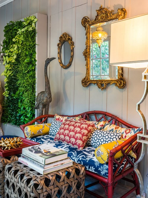Colorful seating area with red bamboo loveseat, patterned cushions, books, woven ottomans, gold mirrors, standing crane sculpture, wall lamp, and a vertical indoor plant display on a panelled wall.